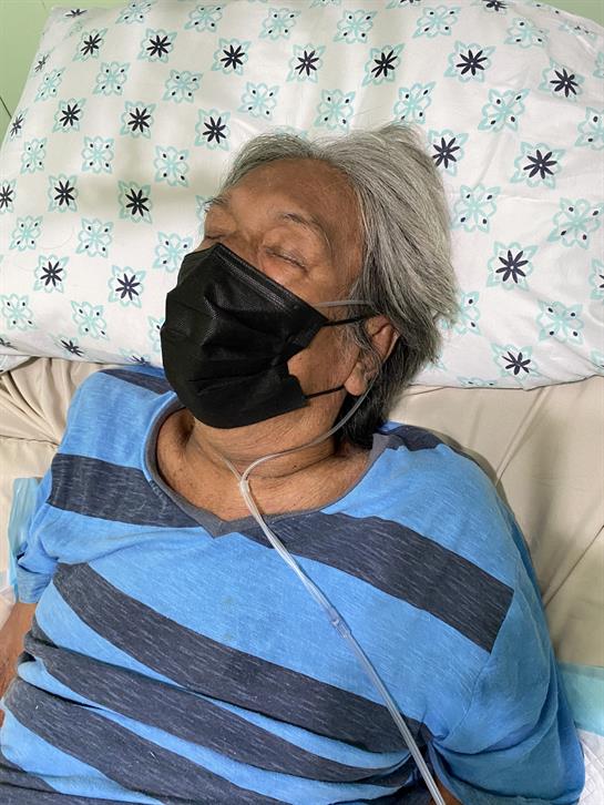 Elderly individual appears to be resting peacefully in a hospital bed while wearing a mask.