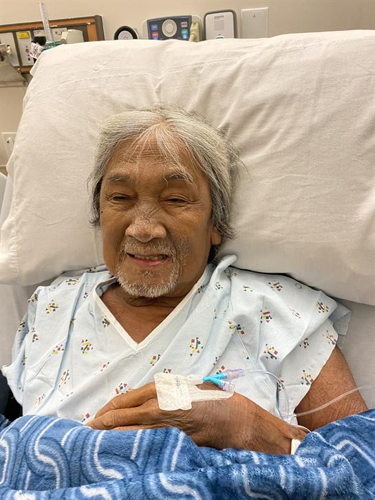 An elderly man in a hospital bed smiles warmly while wearing a medical gown.