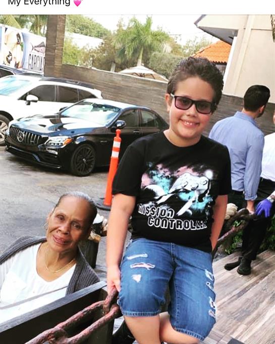 A young boy smiles while sitting next to an elderly woman, surrounded by people and parked cars.