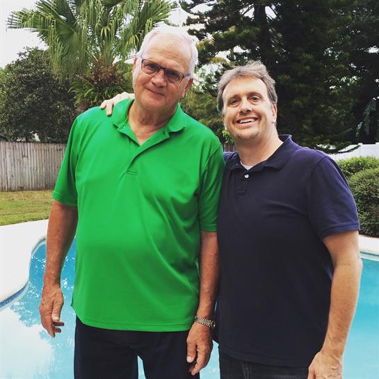Two friends smile together by a clear pool surrounded by palm trees, enjoying their time outdoors.