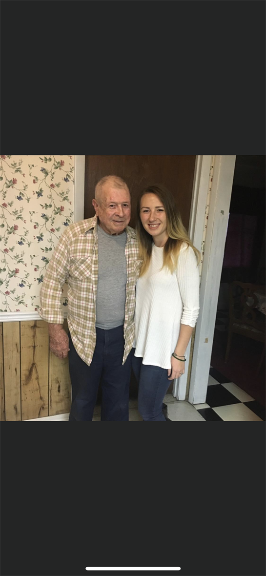 Elderly man smiles while standing next to young woman in a warm and inviting home atmosphere.