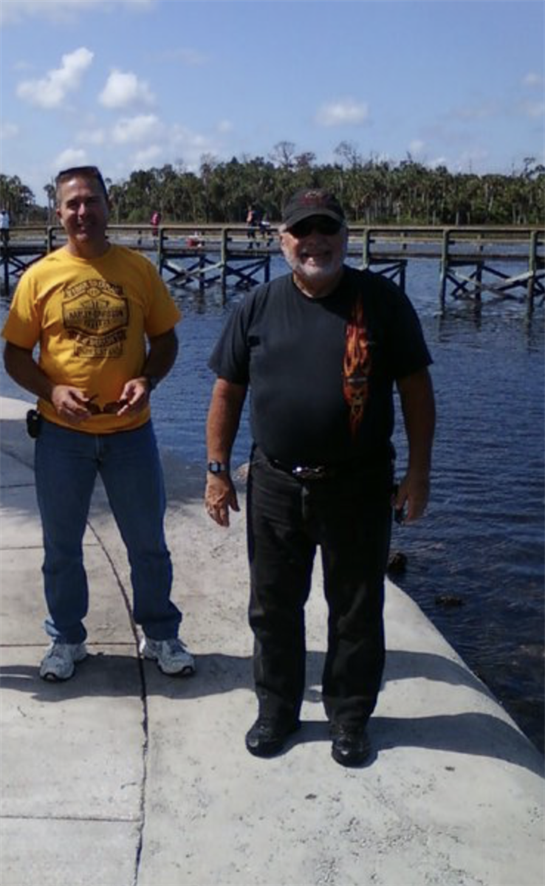 Two friends stand on a dock by the water, smiling and embracing a beautiful day together.