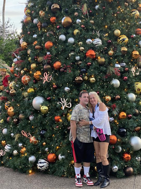 A couple poses happily together by a large, festive Christmas tree adorned with colorful ornaments.