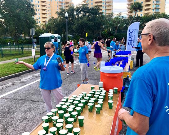 Volunteers are preparing and distributing water cups to runners at a community event in a park.