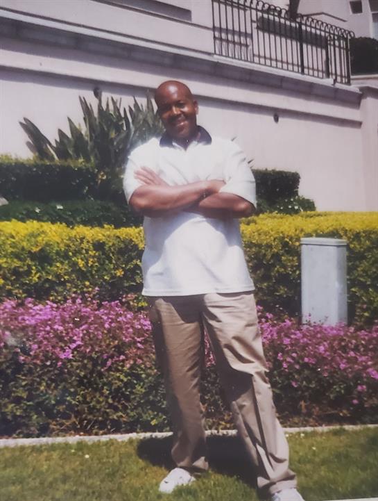 A man stands with arms crossed amidst colorful flowers in a well-kept garden.