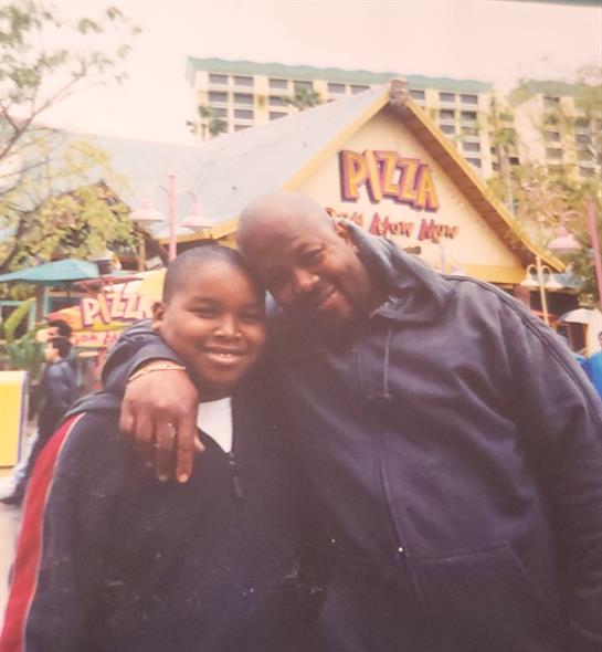 A joyful boy and an adult stand close together, smiling widely by a colorful pizza stand.
