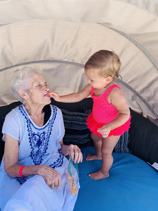 A young girl in a red outfit playfully feeds an elderly woman while sitting together.