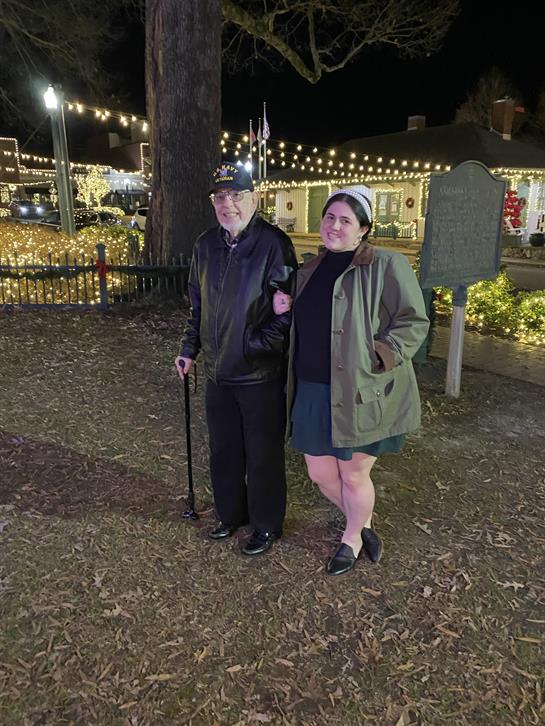 Elderly man and woman walk together under festive lights in a winter park setting.