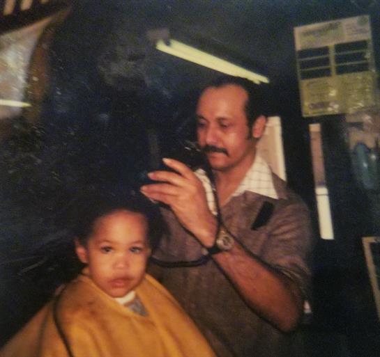 A young child sits in a barber chair, looking slightly serious while getting a haircut.