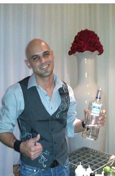 A man holds a liquor bottle, posing confidently next to a large vase with red flowers.
