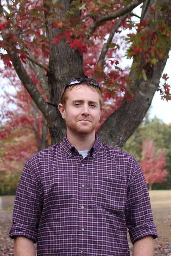 A young man poses confidently in a park surrounded by vibrant red and orange autumn leaves.