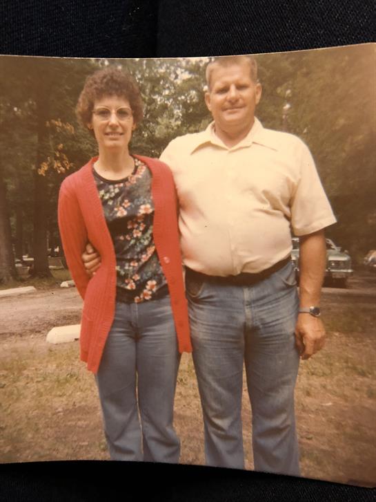 Two individuals stand closely together in a park, smiling at the camera in casual clothing.