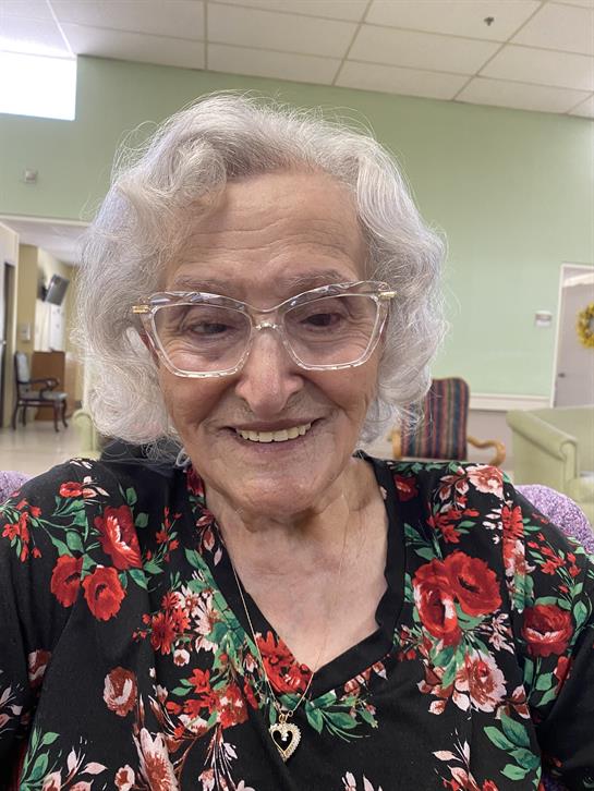 Smiling elderly woman with glasses wears a floral dress in a well-lit community space.