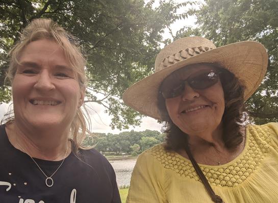Two women smile by the sunny lake, wearing stylish hats with trees behind them.