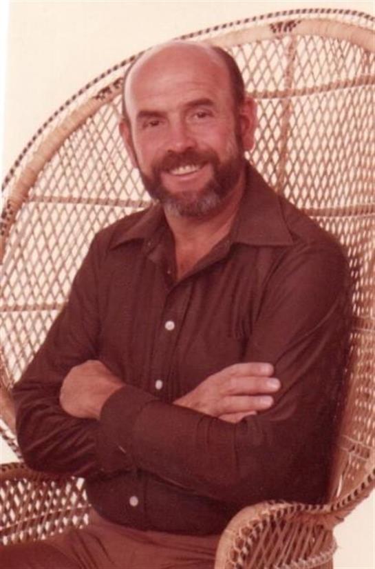 A bearded man smiles confidently in a woven chair, radiating relaxation.