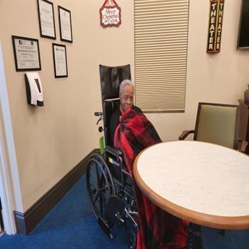An elderly person in a wheelchair sits comfortably, wrapped in a warm blanket in a cozy room.