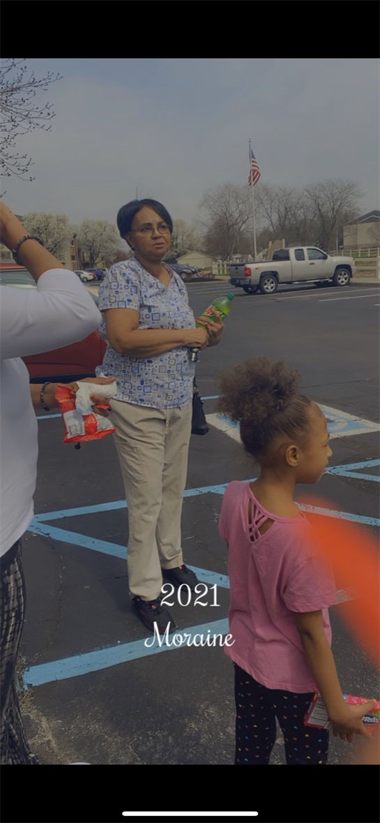 Two adults and a child stand in a parking lot enjoying conversation and refreshments on a sunny day.