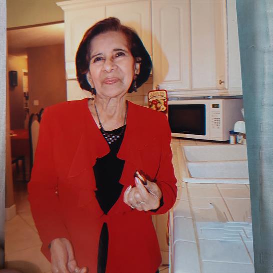 An elderly woman stands in a cozy kitchen, smiling while preparing food and wearing a red jacket.