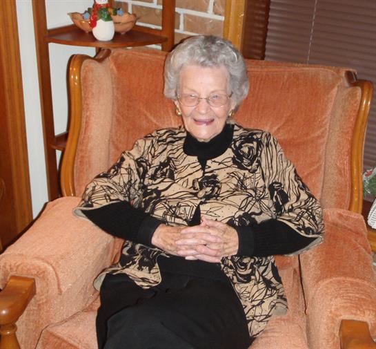 An elderly woman smiles warmly while seated in an armchair at a relaxed indoor gathering.
