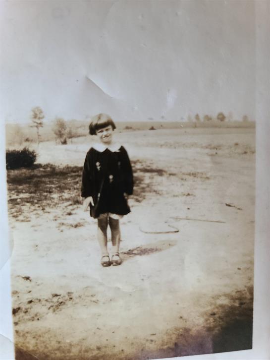 A young girl poses on a dirt path wearing a dark dress and sandals in a vast rural landscape.