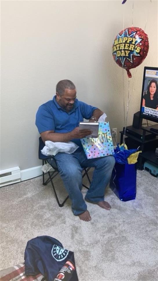 Man sits in a chair as he unwraps a colorful gift while watching television in a cozy setting.