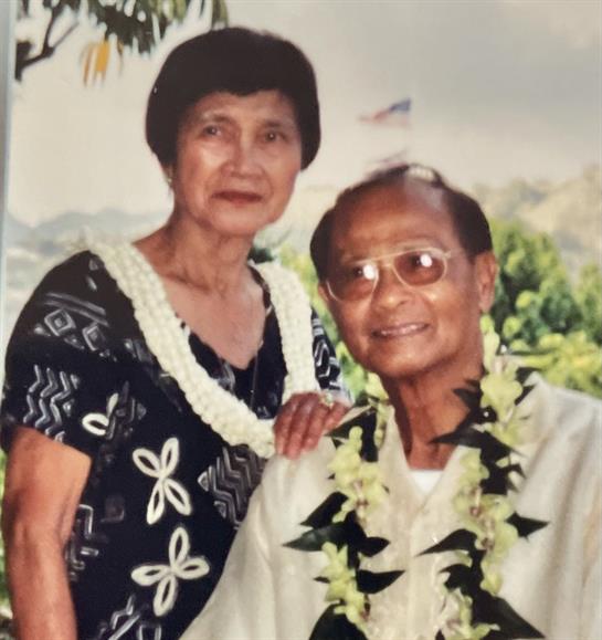 A happy couple in leis poses outdoors amid lush greenery on a sunny day.