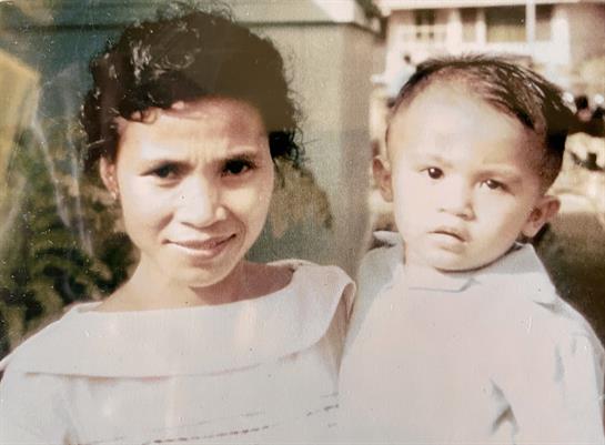 A woman smiles lovingly while holding a young child in her arms in a sunny outdoor setting.