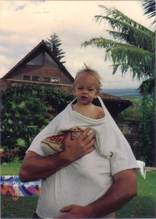 Parent holds child in shirt while posing in a garden with greenery and a house backdrop.