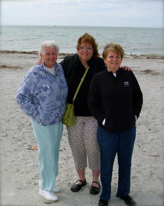 Three friends stand together on a sandy beach, smiling at the ocean as gentle waves lap nearby.