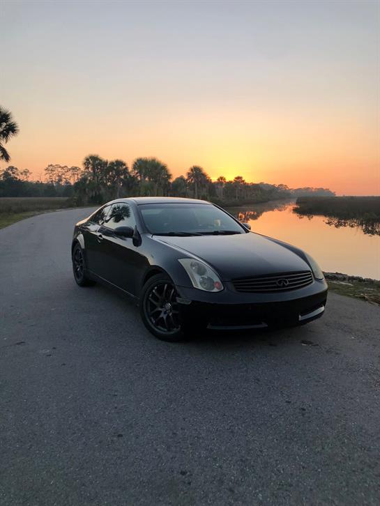 A sleek black sports car is parked by the water, reflecting the beautiful sunset landscape.