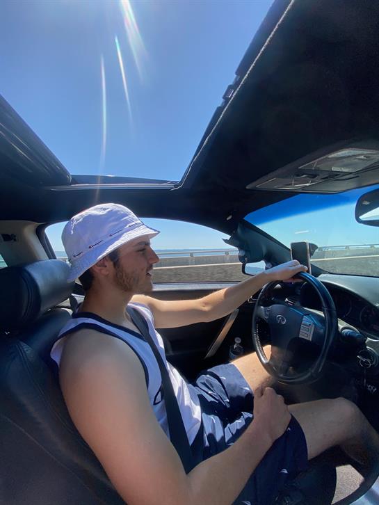 A young man enjoys driving a convertible car along a coastal highway under a clear blue sky.