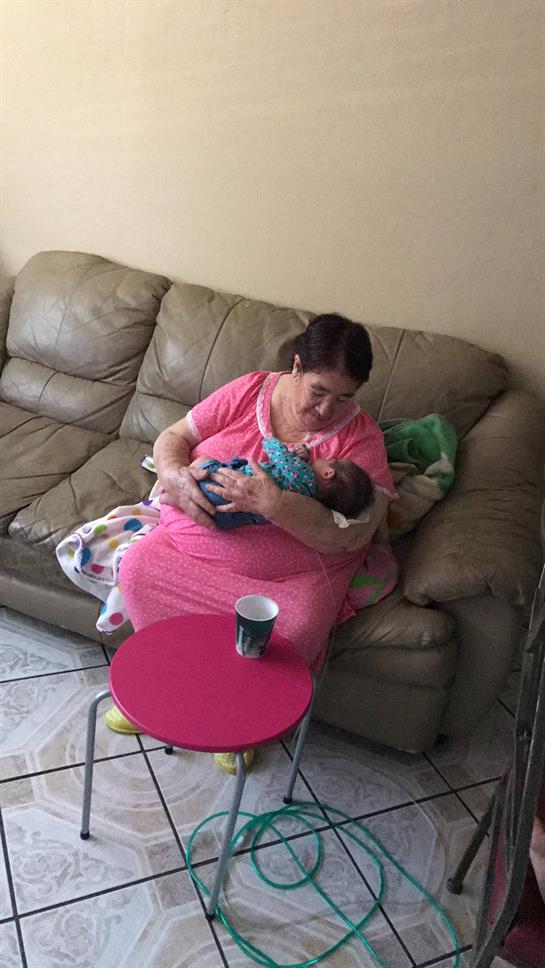 A woman sits on a couch, gently cradling a baby while wearing a pink dress and looking content.
