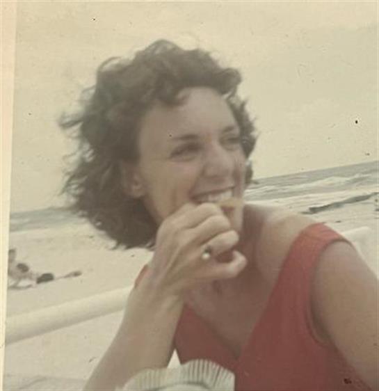 A woman smiles brightly while eating a snack at the beach during a sunny day, embodying happiness.