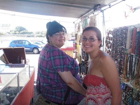 Two women are playfully interacting at a jewelry vendor stall during a sunny market day.
