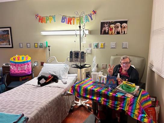 Friends gather to celebrate a birthday in a hospital room decorated with colorful banners and gifts.