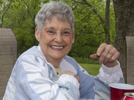 Elderly woman sits in a park, smiling widely while engaging in conversation with friends.