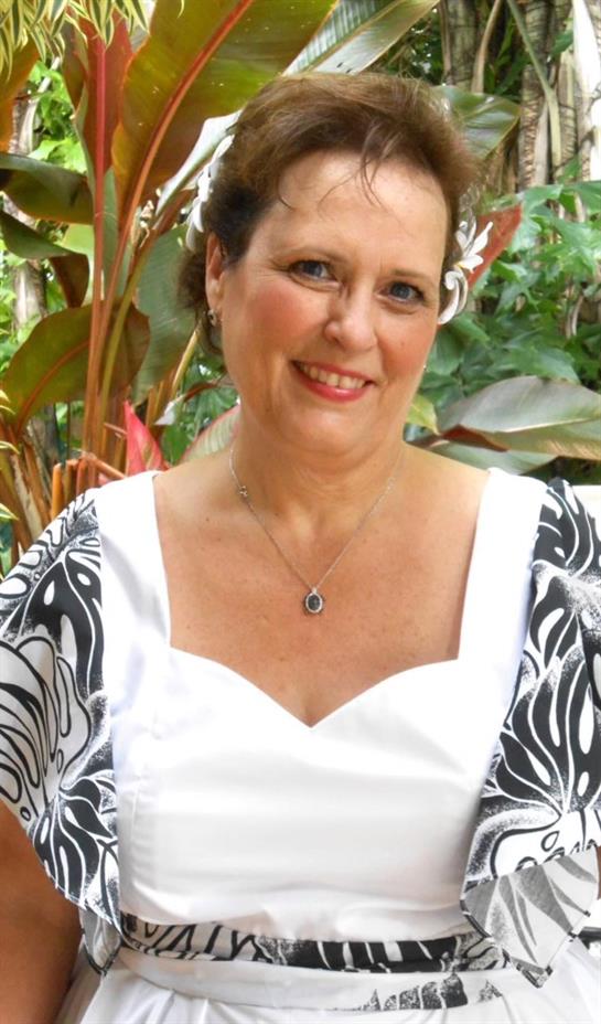 A woman smiles in a white dress adorned with floral details surrounded by greenery.