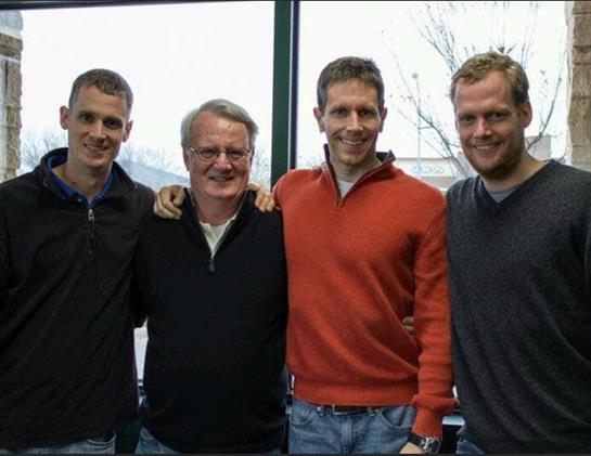 Four men stand together, smiling and posing with arms around each other in a cozy indoor setting.