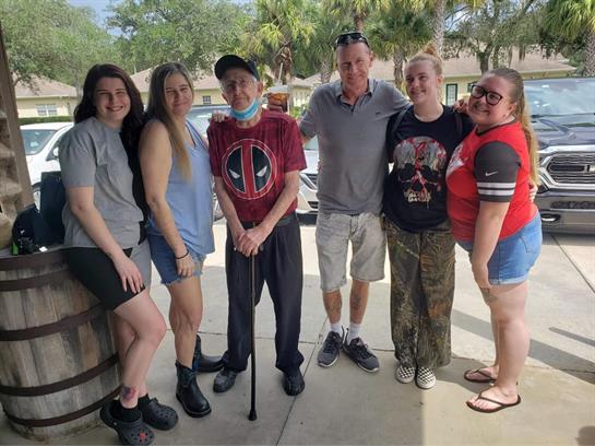 A group stands together smiling outdoors, with an elderly man holding a cane and wearing a mask.