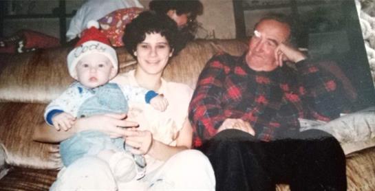 A woman holds a baby wearing a holiday hat while an elderly man looks on, relaxed on a couch.