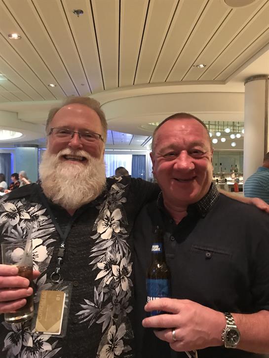 Two smiling men hold drinks and share a moment in a vibrant social setting with friends.