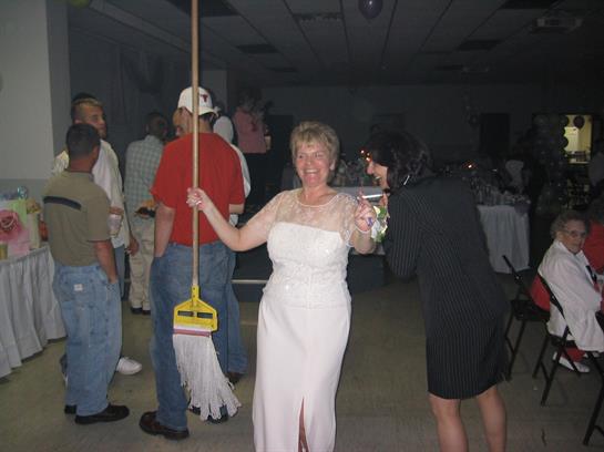 A woman in a white dress joyfully poses with a broom at a festive gathering.