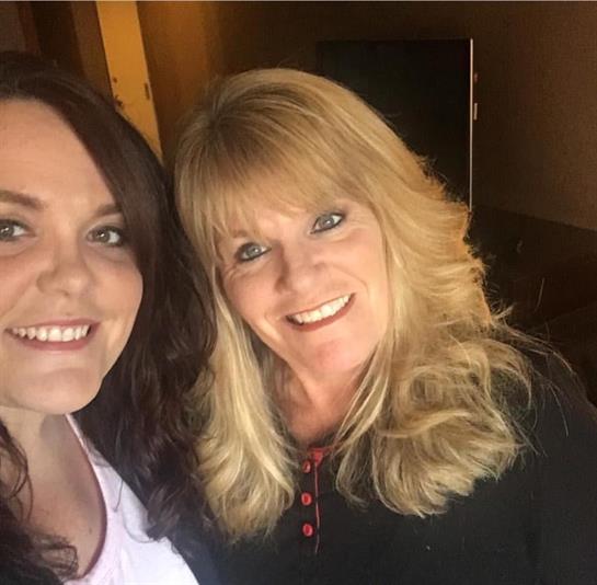 Two women share a joyful moment together, smiling for a selfie indoors with warm lighting.