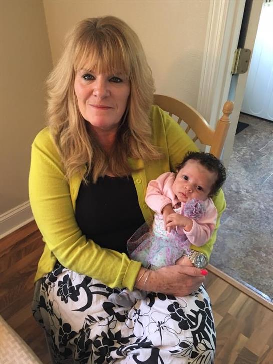 A smiling woman with blonde hair sits on a chair, holding a baby in her arms, both appearing happy.