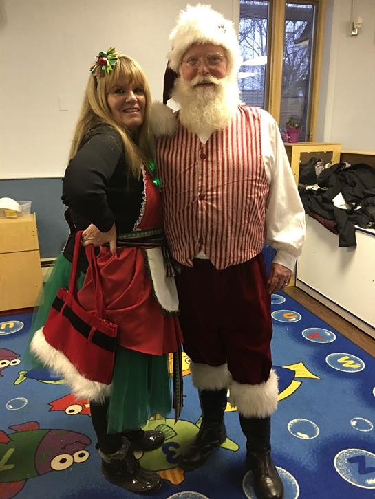 Santa and a helper cheerfully pose in a lively indoor winter wonderland.