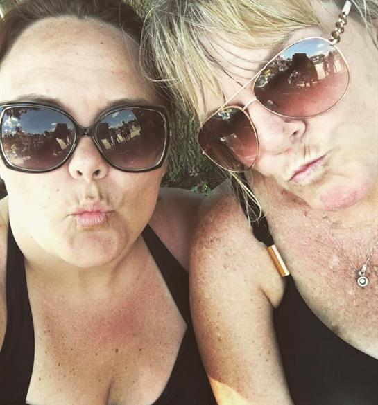 Two friends happily pose outdoors in sunglasses, enjoying the sunny day.