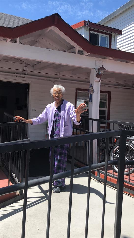 An elderly woman is smiling and waving from her porch, enjoying a lovely sunny day outside.
