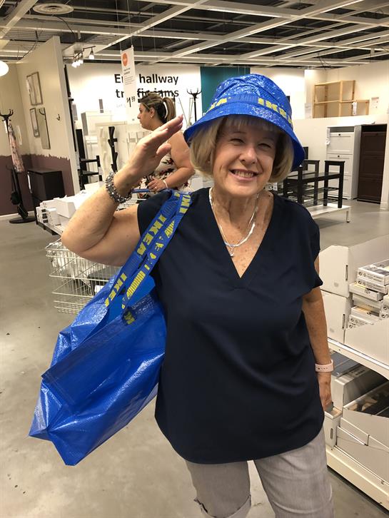 Cheerful older woman waves while shopping indoors with a blue bag and hat on.