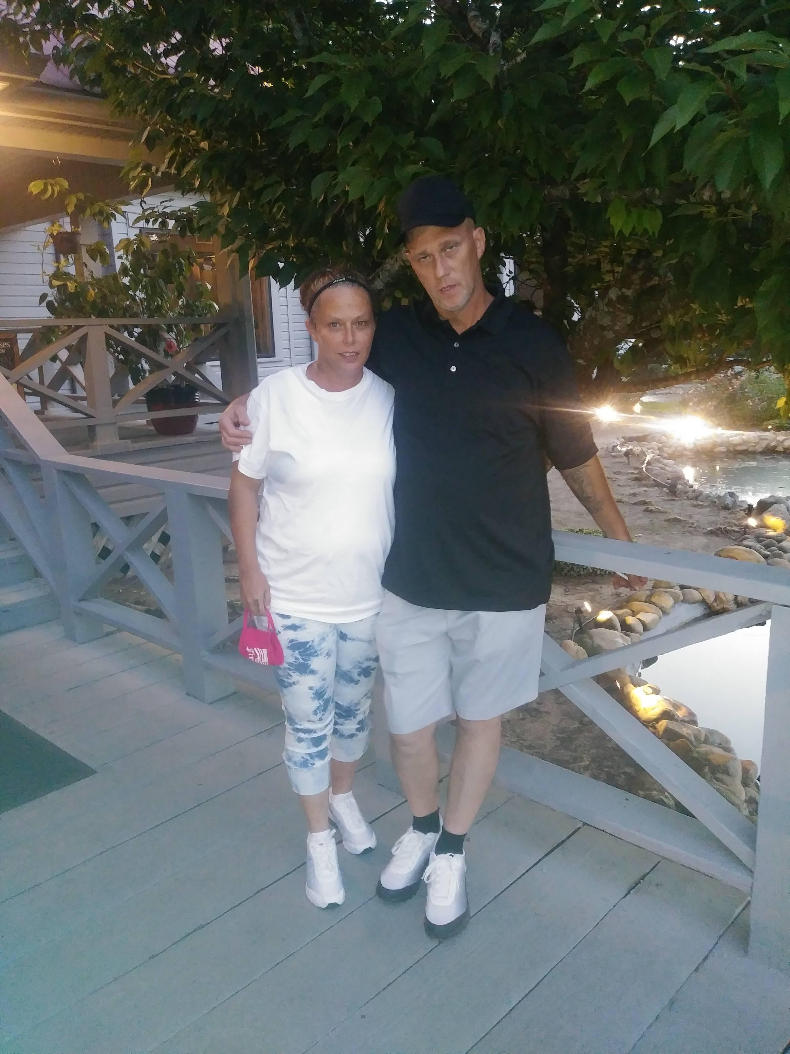 Two people stand close together on a pathway near the water, smiling as the sun sets behind them.