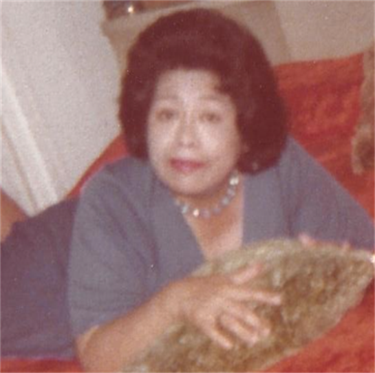An elderly woman lies on a couch, gently holding a decorative pillow while smiling warmly.